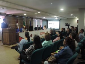 Pessoas reunidas em um auditório. O Procurador Geral de Justiça fala ao microfone, em pé, diante de uma tribuna.