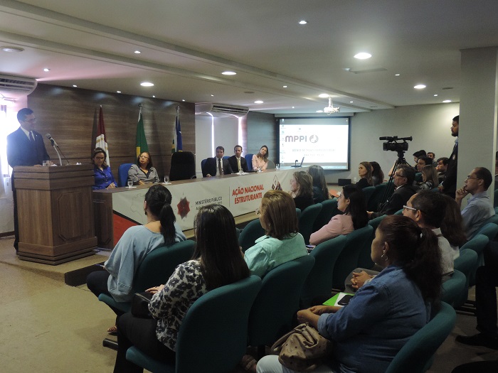 Pessoas reunidas em um auditório. O Procurador Geral de Justiça fala ao microfone, em pé, diante de uma tribuna.