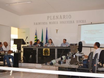 A Promotora de Justiça está sentada no centro de uma mesa, em um plenário, falando ao microfone a um público.