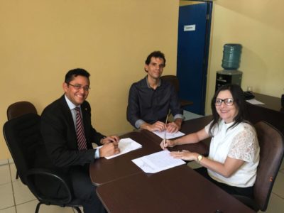 Dois homens e uma mulher sentados junto a uma mesa, sorrindo. Um deles está assinando um documento.