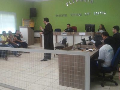 Promotor de Justiça, em pé, de toga, fala para os membros do Conselho de Sentença, que estão sentados à frente dele.