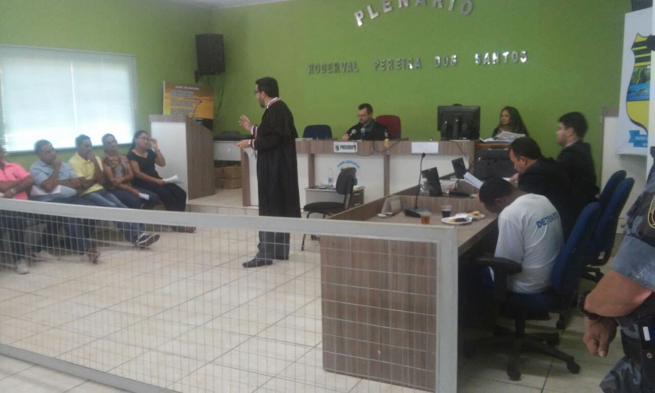 Promotor de Justiça, em pé, de toga, fala para os membros do Conselho de Sentença, que estão sentados à frente dele.