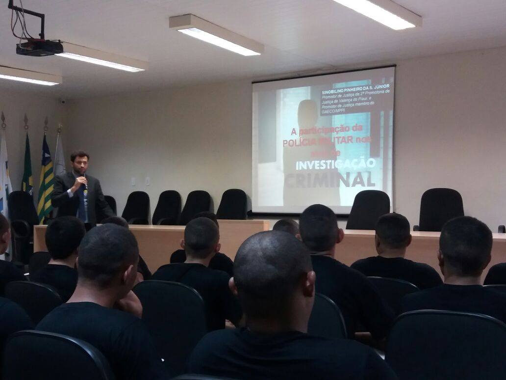 Promotor de Justiça em pé no auditório, ladeado por vários policiais militares fardados.