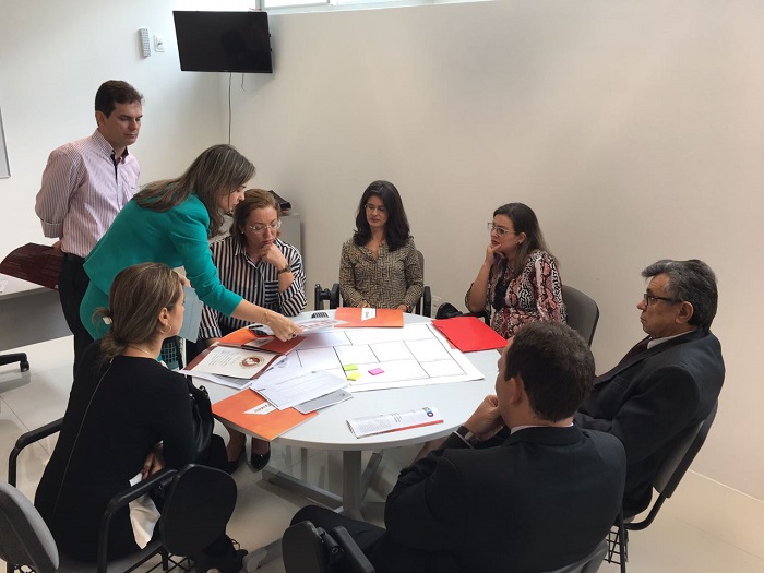 Pessoa de pé dando instruções para outras que estão sentadas em volta de uma mesa 