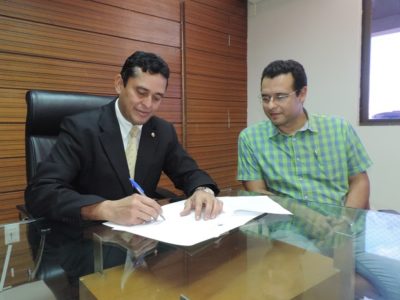 Duas pessoas sentadas na mesa, uma assinando um documento, enquanto o outro observa