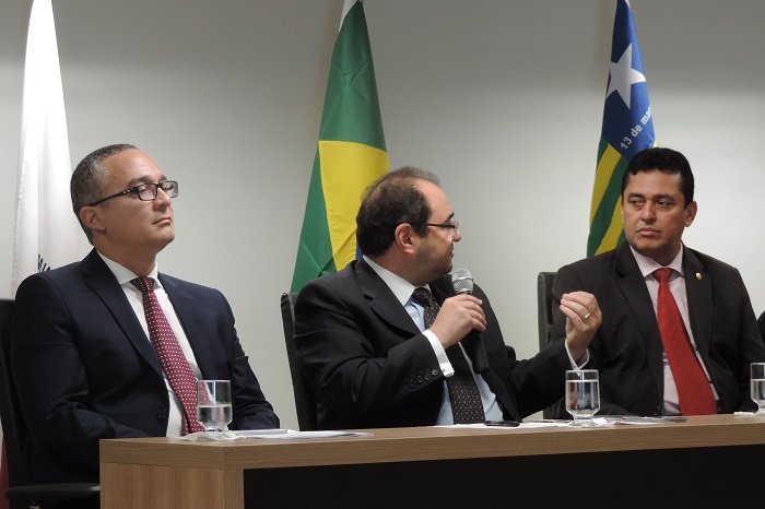 Três homens sentados, dois se olham enquanto fala