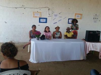 Pessoas sentadas em cadeiras, em um pátio. A foto mostra uma mesa de honra com quatro pessoas, sendo que uma delas é a Promotora de Justiça Leida Diniz.