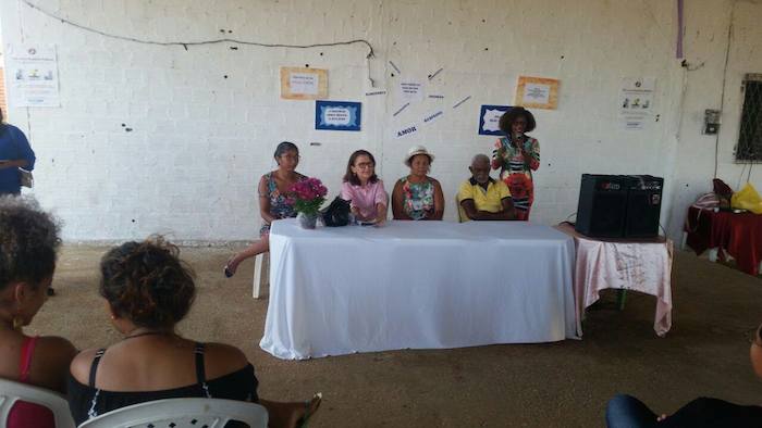 Pessoas sentadas em cadeiras, em um pátio. A foto mostra uma mesa de honra com quatro pessoas, sendo que uma delas é a Promotora de Justiça Leida Diniz.
