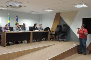 Mulher de pé com microfone nas mãos, enquanto outras pessoas a observam