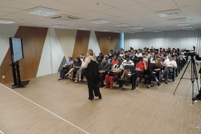 Pessoas de sentadas em auditório e um mulher de pé com microfone na mão 