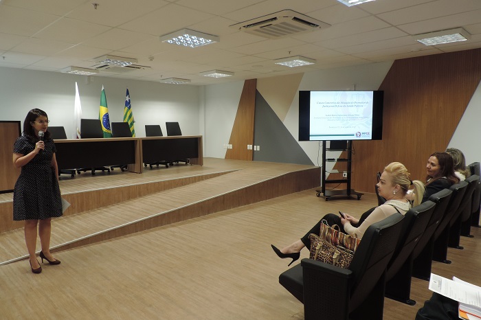 Mulher de pé com microfone nas mãos, enquanto as demais pessoas estão sentadas