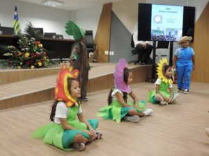 Crianças vestidas de flor e de árvore se apresentam em um auditório.