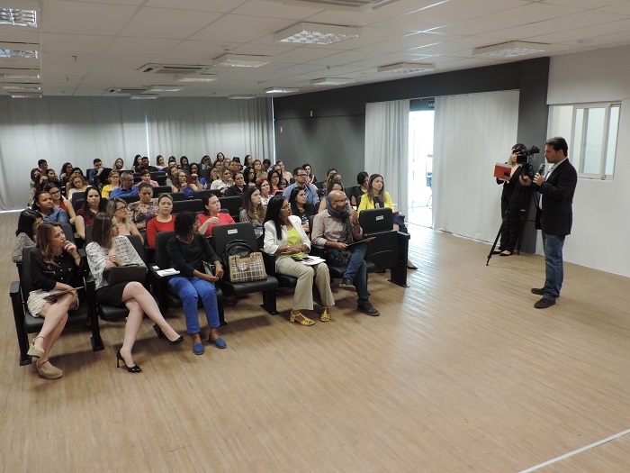 Homem de pé com microfone na mão, enquanto as demais pessoas sentadas e o observam 