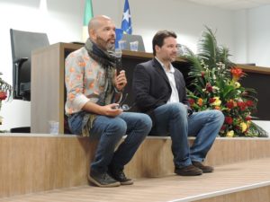 Dois homens sentados. Um deles segura um microfone entre as mãos