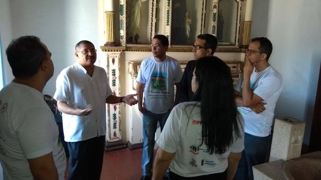 Um padre fala a cinco pessoas que estão em círculo, dentro de uma igreja.