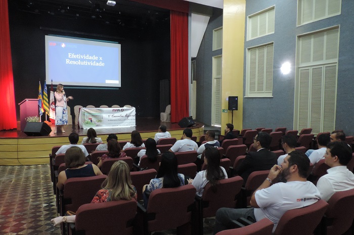 Um auditório com várias pessoas sentadas. Ao fundo, uma palestrante fala no palco.