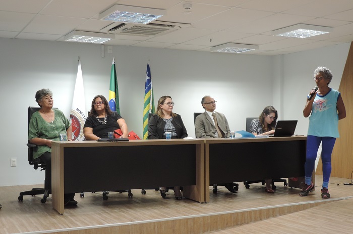 Pessoas em mesa de honra, em auditório. Uma senhora idosa, de pé, fala ao microfone.