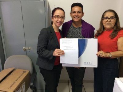 Três pessoas de pé sorrindo para fotografia. As três seguram um papel e uma caixa com o computador