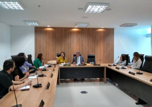 Pessoas sentadas em volta de uma mesa. Enquanto uma mulher gesticula falando e os demais observam