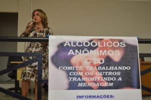 Mulher de pé com microfone na mão, e um cartaz na frente com informações sobre alcoolismo