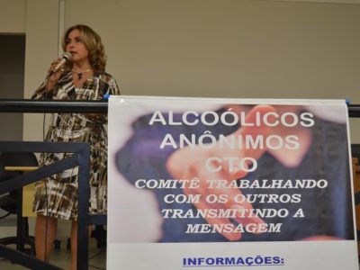 Mulher de pé com microfone na mão, e um cartaz na frente com informações sobre alcoolismo