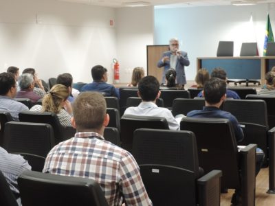 Pessoas sentadas num auditório observando um homem de pé com microfone nas mãos