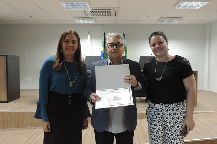Três pessoas de pé, ao centro um homem segurando um certificado 