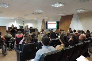 Auditório lotado. Na foto, nota-se alguns cadeirantes. Na frente, uma palestrante e uma intérprete de LIBRAS.