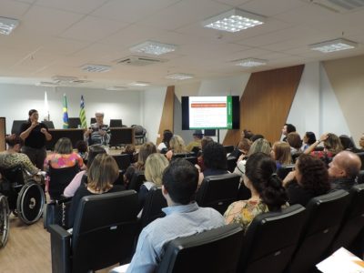 Auditório lotado. Na foto, nota-se alguns cadeirantes. Na frente, uma palestrante e uma intérprete de LIBRAS.