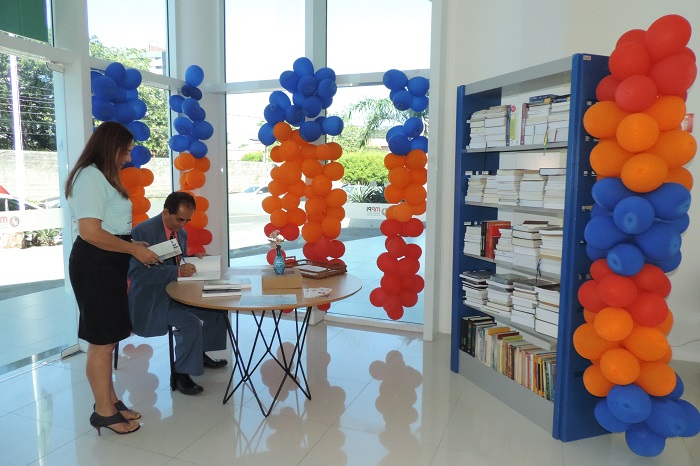Duas pessoas, uma sentada e outras de pé. Ao fundo há uma estante de livros e balões decorativos 
