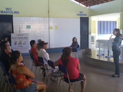 Procuradora de Justiça fala para algumas pessoas sentadas em cadeiras, no pátio de uma escola.