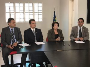 Quatro pessoas em uma mesa. O Procurador-Geral está com uma caneta na mão, prestes a assinar um documento.