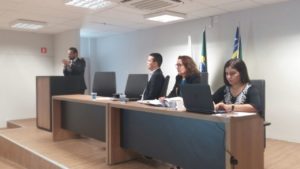 Promotora de Justiça e mais duas pessoas sentadas à mesa principal de um auditório. Um homem fala junto à tribuna, de pé.