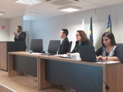 Promotora de Justiça e mais duas pessoas sentadas à mesa principal de um auditório. Um homem fala junto à tribuna, de pé.
