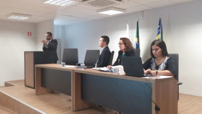 Promotora de Justiça e mais duas pessoas sentadas à mesa principal de um auditório. Um homem fala junto à tribuna, de pé.