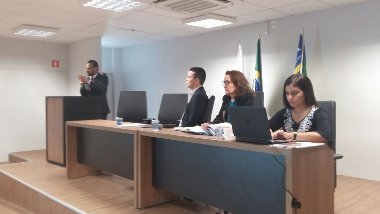 Promotora de Justiça e mais duas pessoas sentadas à mesa principal de um auditório. Um homem fala junto à tribuna, de pé.
