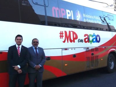 Dois homens de terno posam de pé. Atrás dele, está um grande ônibus em perspectiva lateral, com adesivos em que se lê