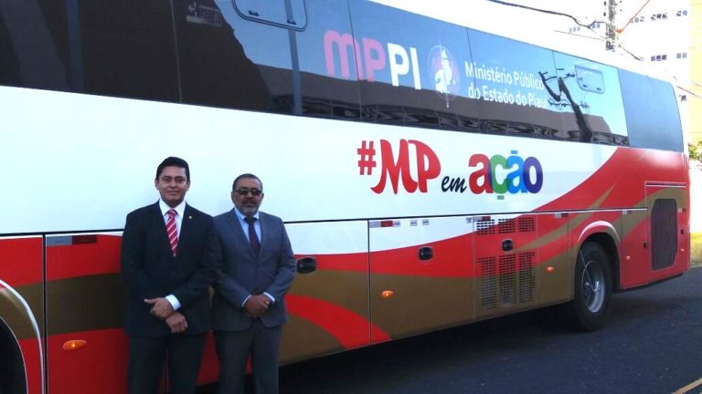 Dois homens de terno posam de pé. Atrás dele, está um grande ônibus em perspectiva lateral, com adesivos em que se lê