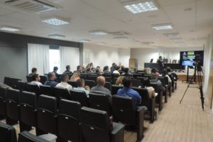 Pessoas sentadas em auditório, ao fundo um homem de pé com mirofone nas mãos