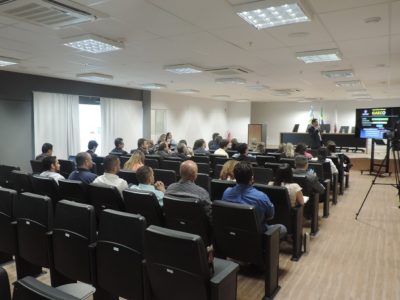 Pessoas sentadas em auditório, ao fundo um homem de pé com mirofone nas mãos