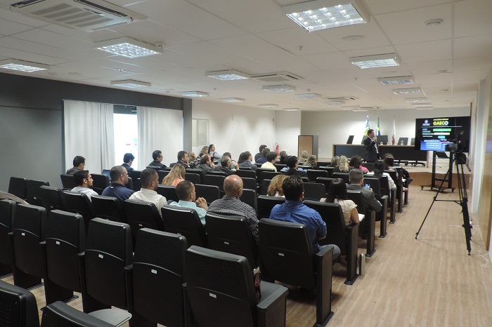 Pessoas sentadas em auditório, ao fundo um homem de pé com mirofone nas mãos