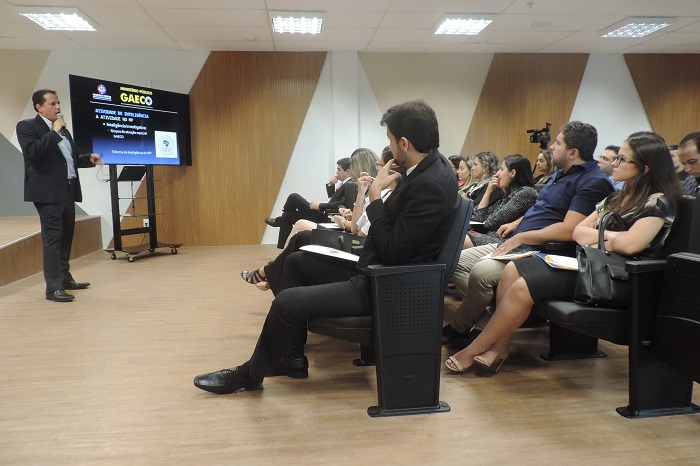 Pessoas sentadas em auditório, ao fundo homem de pé próximo a telão com microfone nas mãos 