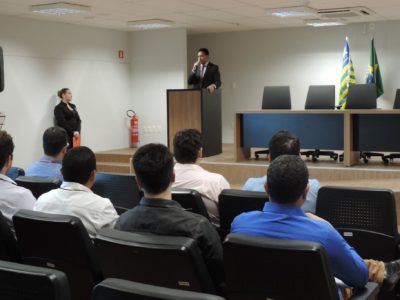 Pessoas sentadas em auditório, ao fundo um homem de pé sgurando um micrfone nas mãos