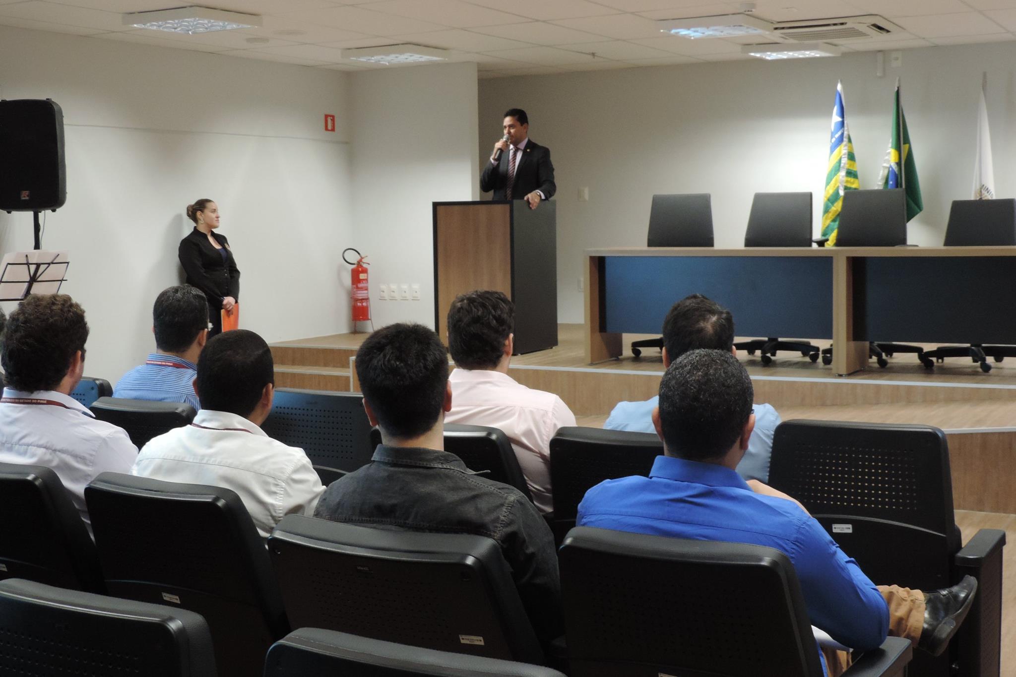 Pessoas sentadas em auditório, ao fundo um homem de pé sgurando um micrfone nas mãos