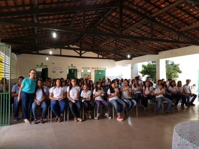 Pessoas sentadas em pátio da escola, uma mulher de pé abraça um dos estudantes