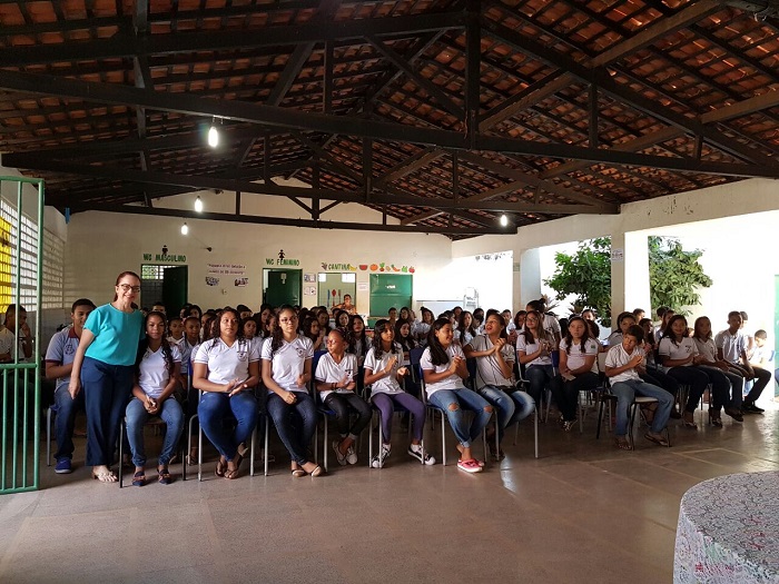 Pessoas sentadas em pátio da escola, uma mulher de pé abraça um dos estudantes