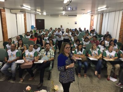 Em primeiro plano uma mulher está de pé segurando um microfone nas mãos, ao fundo pessoas sentadas ouvindo a mulher falando