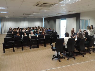 Pessoas sentadas em auditório, ao fundo mulheres sentadas e de pé falando