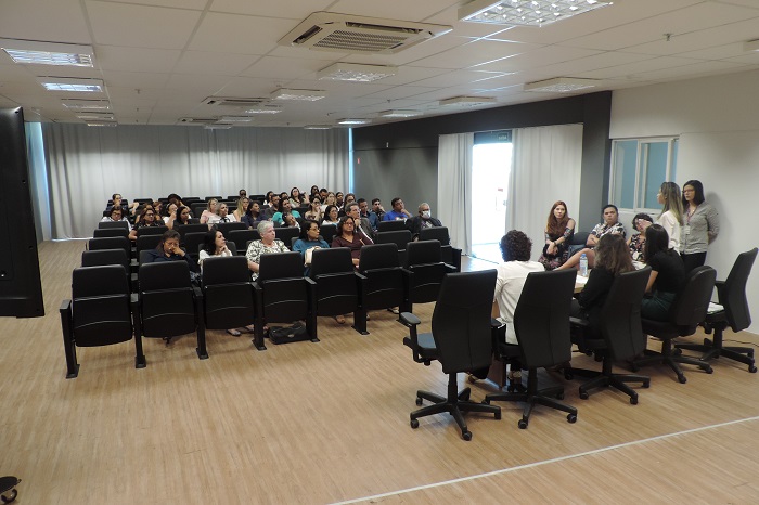 Pessoas sentadas em auditório, ao fundo mulheres sentadas e de pé falando