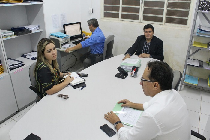 Três pessoas sentadas em volta de uma mesa, ao fundo um homem está sentando em outra mesa manuseando um computador 